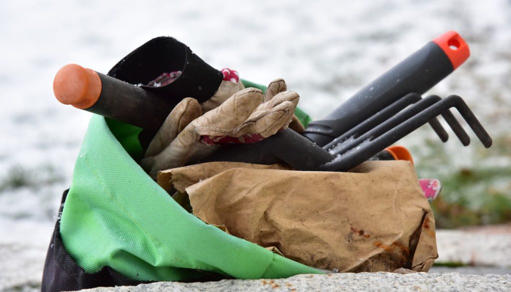A set of gardening tools and gloves in a green bag, placed outdoors on a snowy surface.