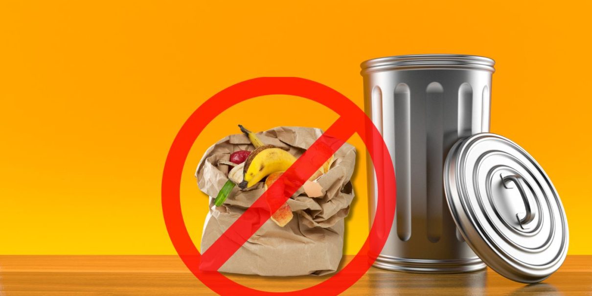 A brown paper bag filled with fruit and vegetable peels crossed out with a red circle, placed next to a metallic trash can against a bright orange background.