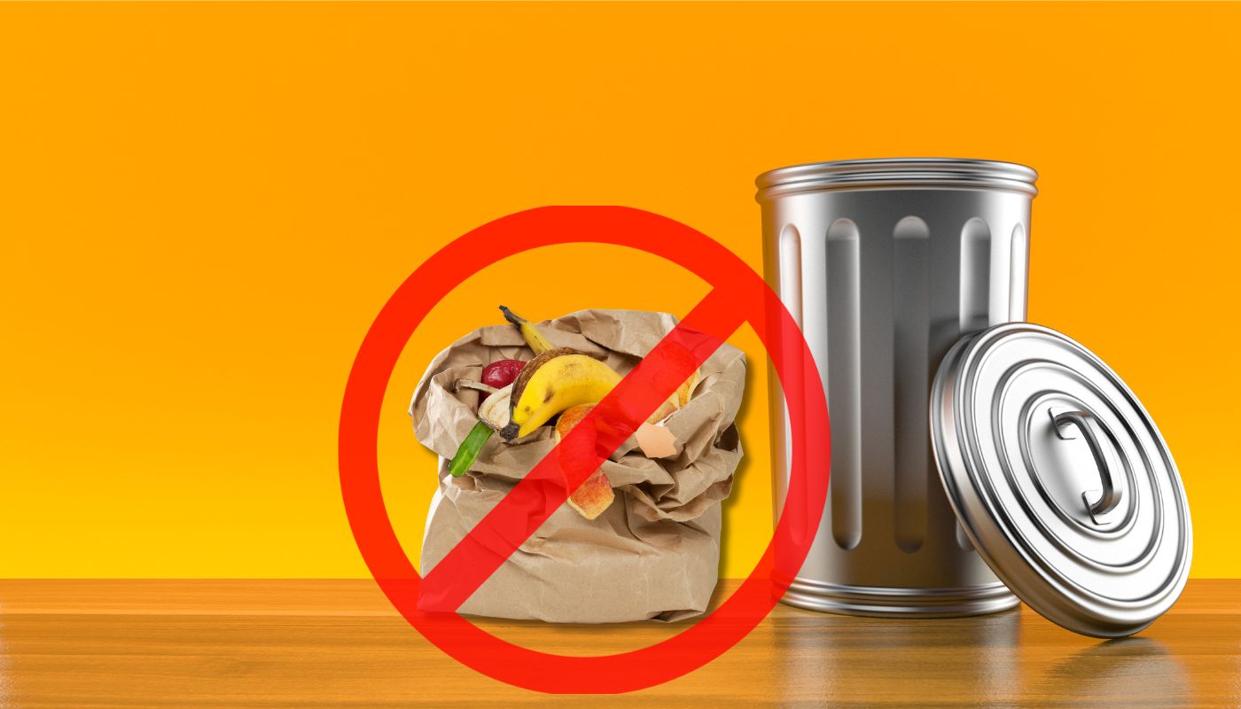 A brown paper bag filled with fruit and vegetable peels crossed out with a red circle, placed next to a metallic trash can against a bright orange background.